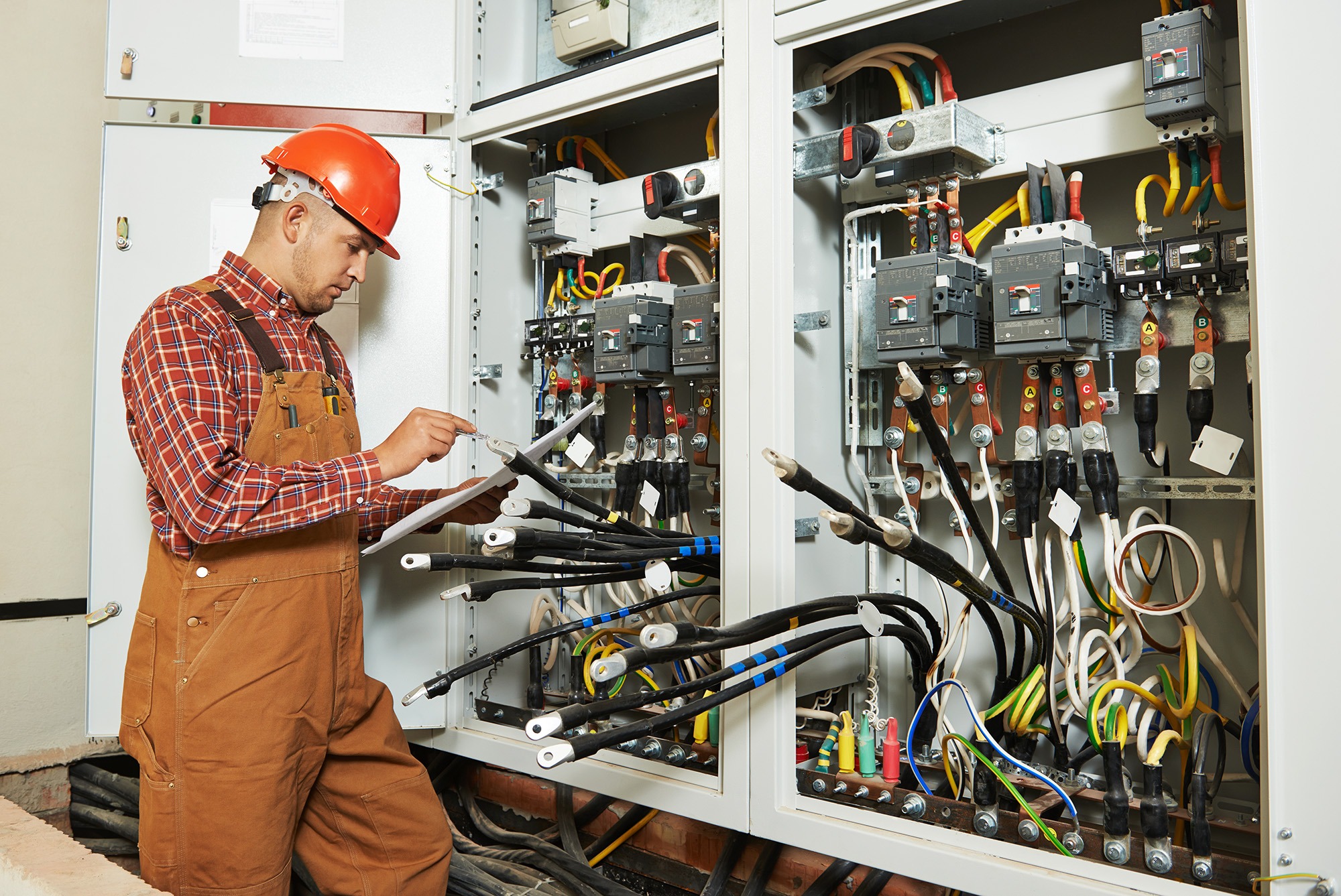An electrician in protective gear inspects wiring inside an electrical panel, taking notes to ensure safety and compliance.