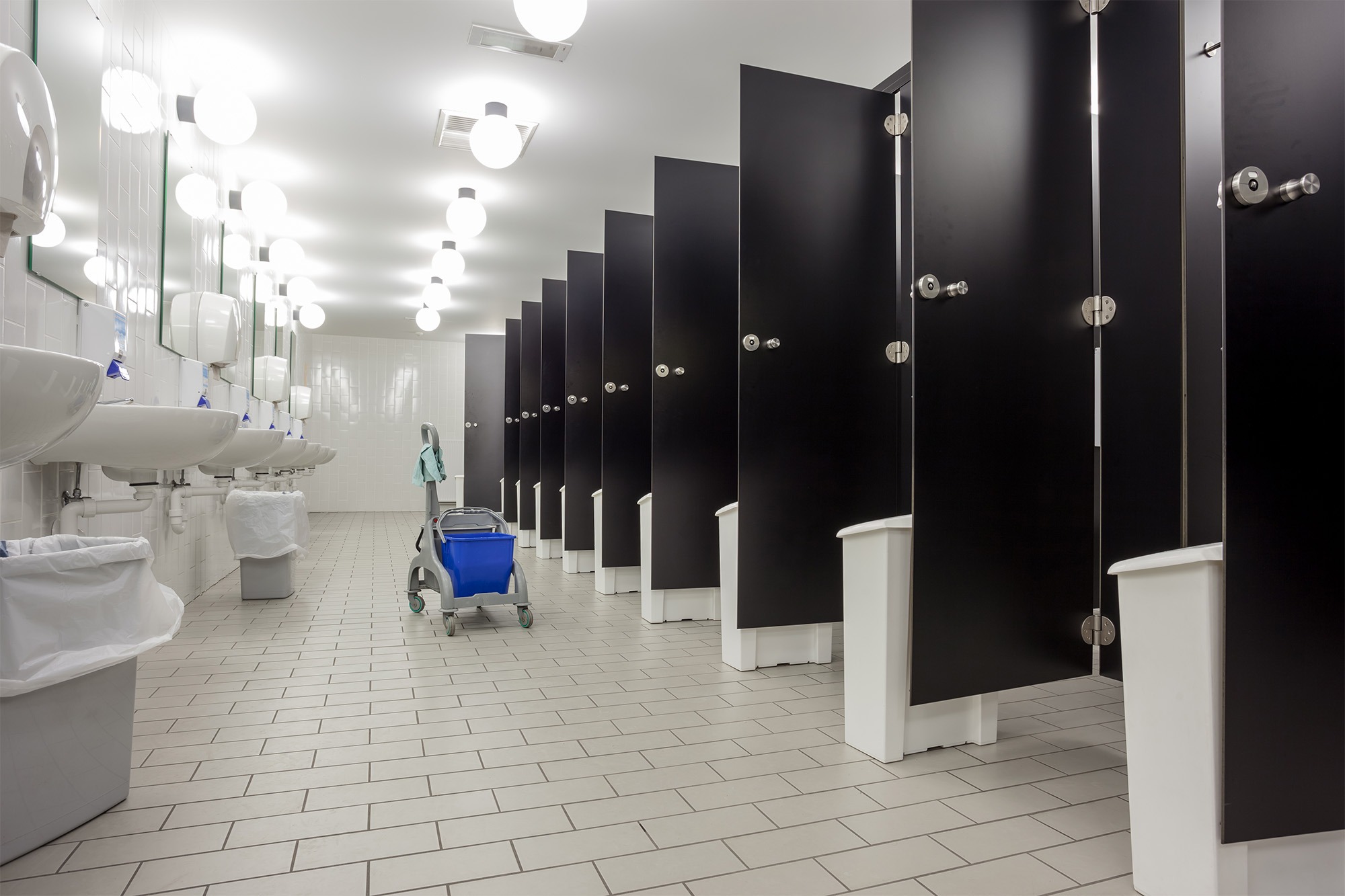 Clean public restroom featuring multiple black stalls, sinks with white basins, and a janitorial cart, emphasizing hygiene and accessibility.