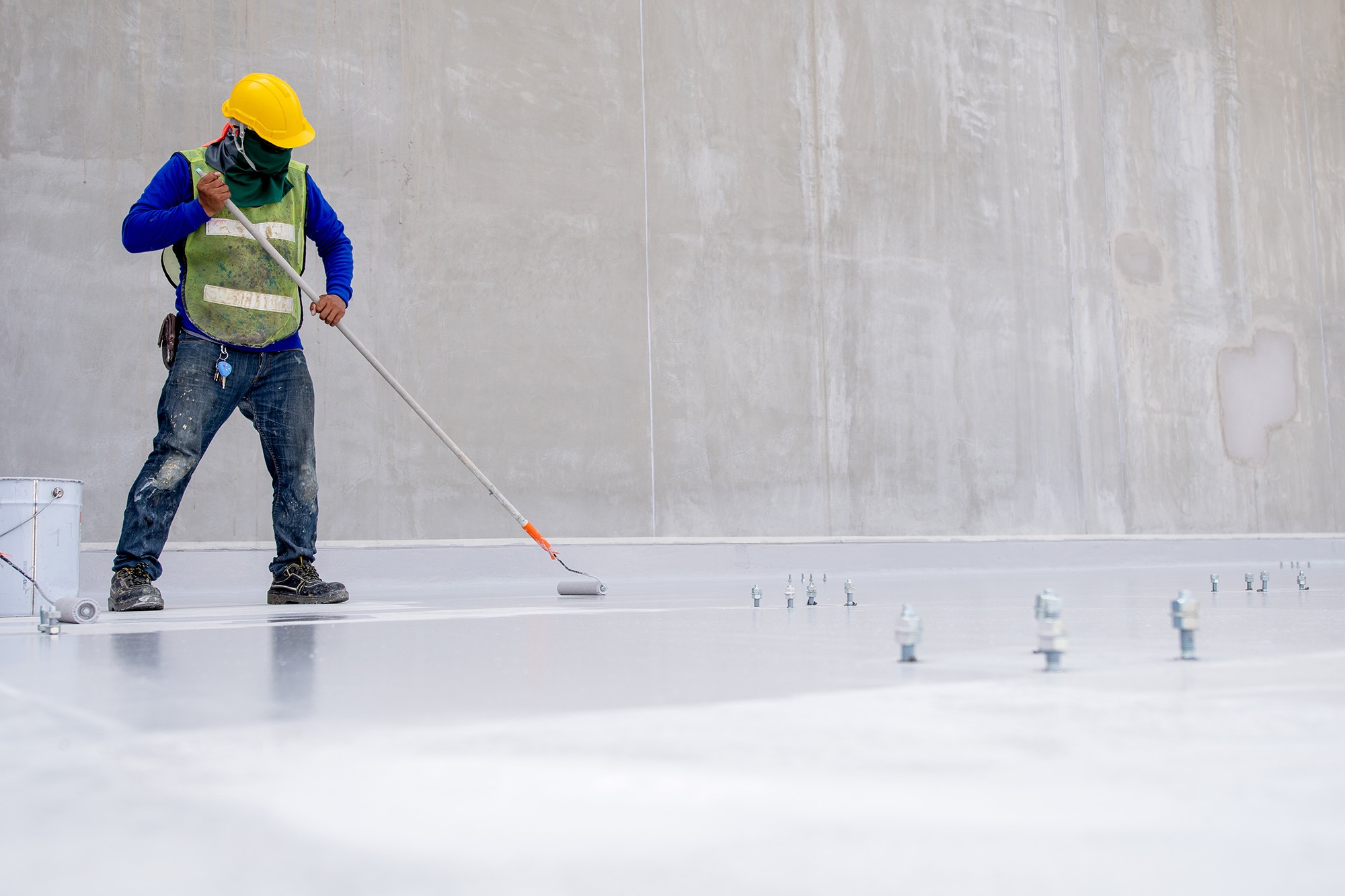 Construction worker in a yellow hard hat and safety vest applies paint to a concrete floor with a roller, showcasing ongoing facility work.