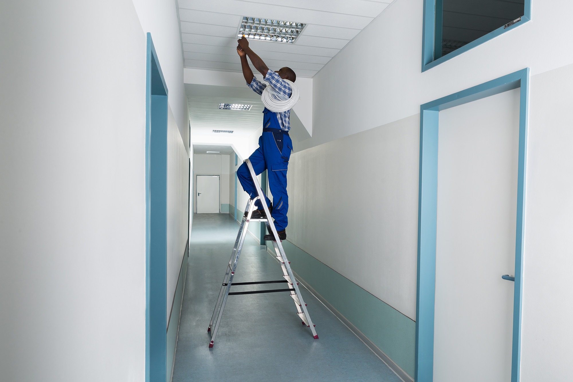 A maintenance worker stands on a ladder adjusting ceiling lights in a well-lit hallway, illustrating the importance of facility upkeep in a professional environment.