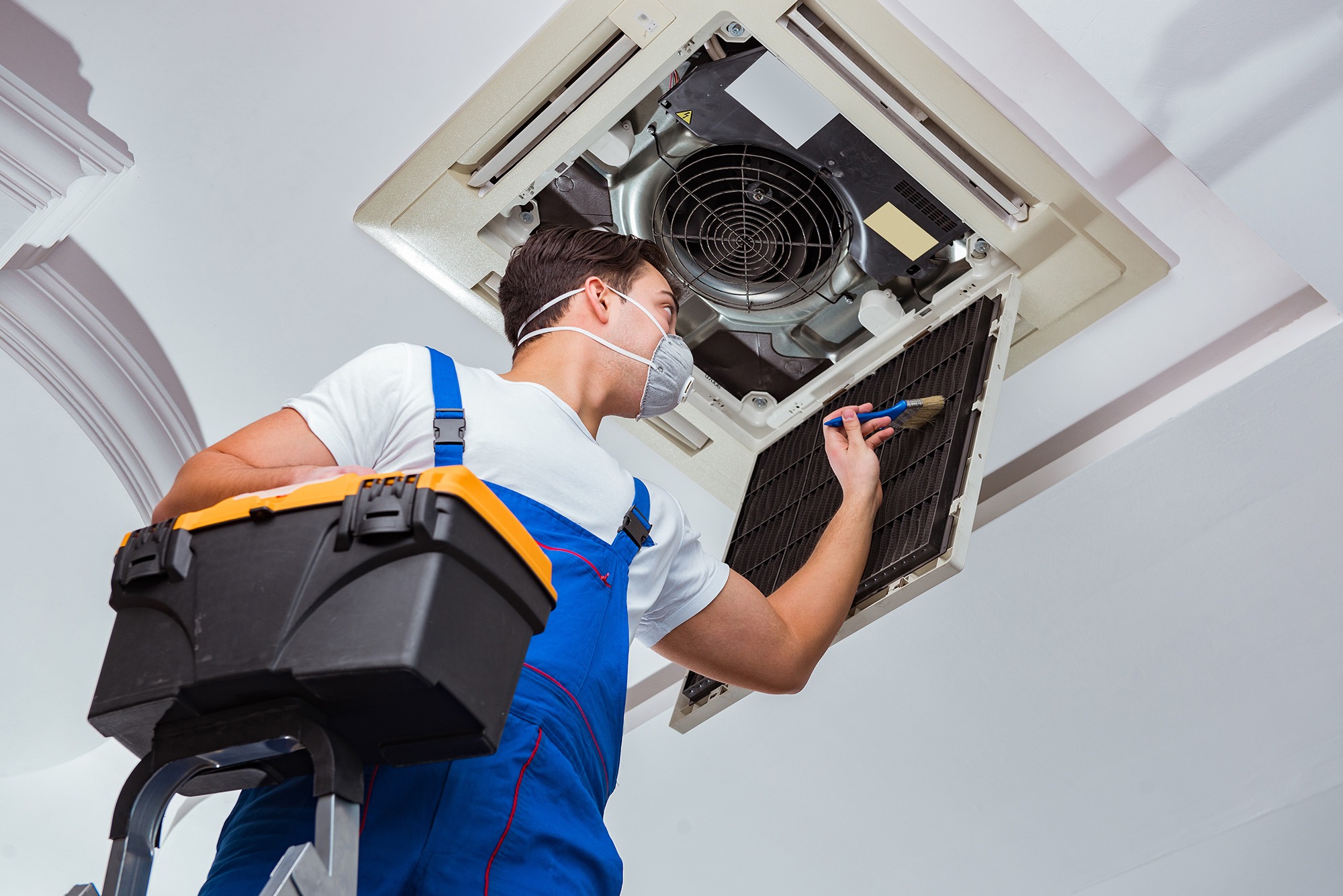Technician in blue overalls performing maintenance on a ceiling air conditioning unit, cleaning filters with a brush while using a toolbox.